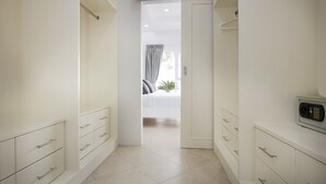 Un couloir étroit, avec des meubles blancs et des tiroirs, qui mène à une pièce où se trouvent un lit et une plante.