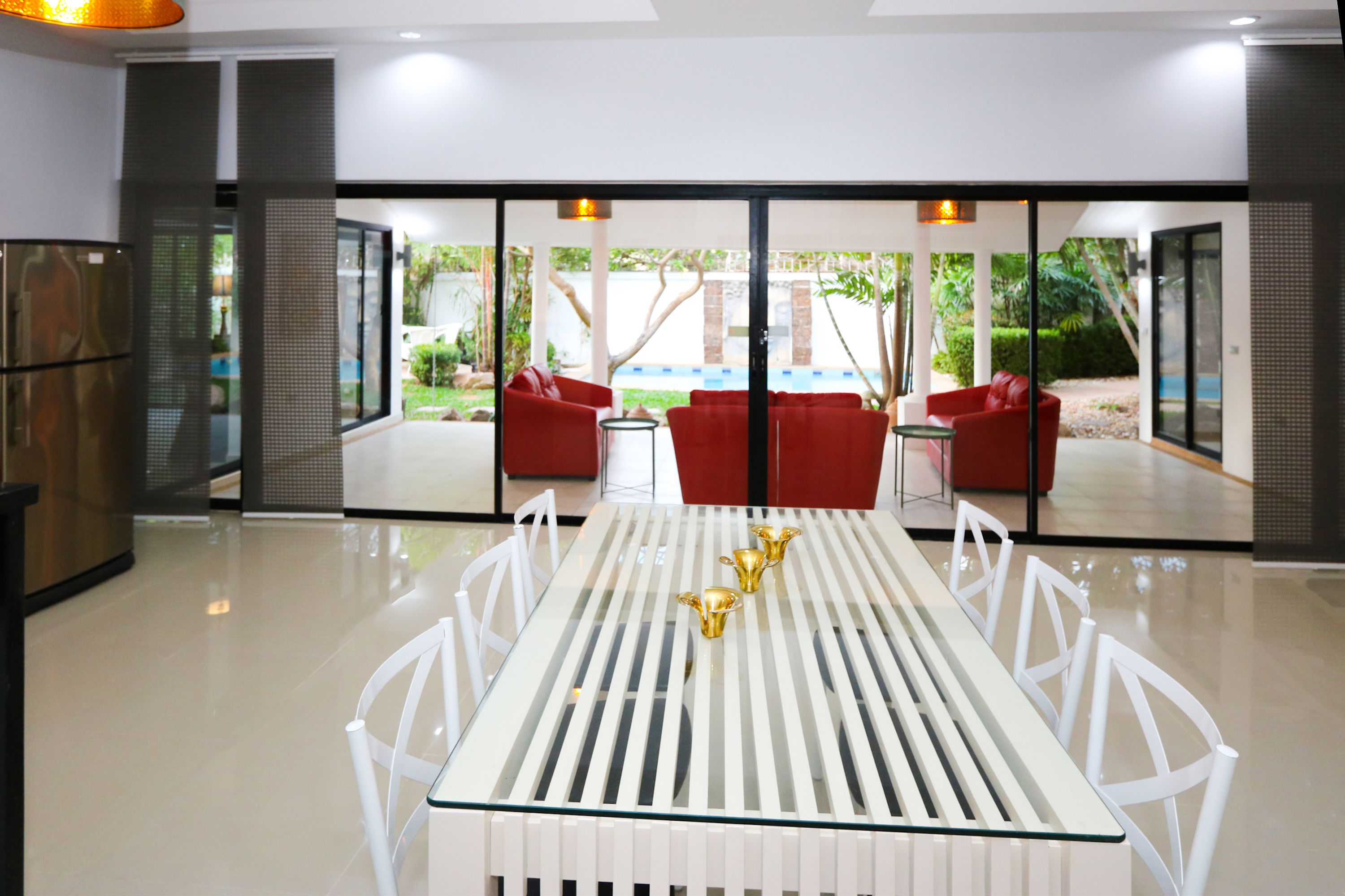A modern dining area with a white table and chairs, a red sofa set, and a glass partition with a view of a pool and greenery.
