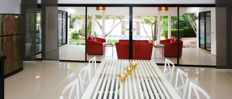 A modern dining area with a white table and chairs, a red sofa set, and a glass partition with a view of a pool and greenery.