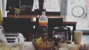 A breakfast table with plates, a basket of bread, a coffee maker, and a window view of a bicycle. - Empathy Guesthouse - Hostel (Daegu)