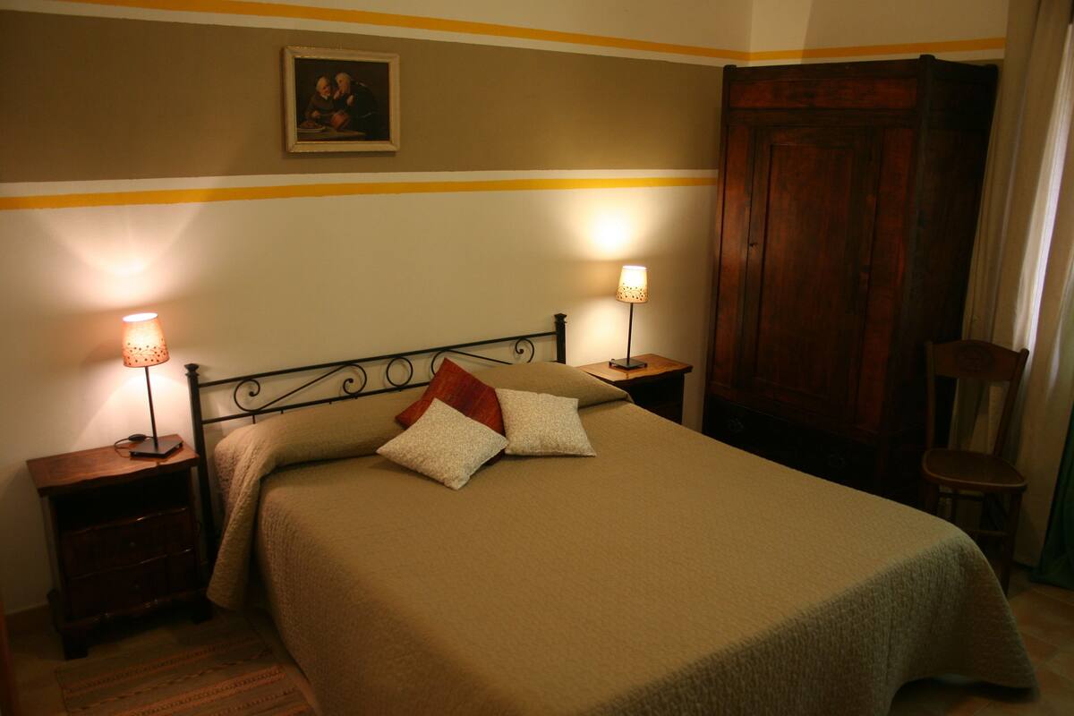 A neatly made bed with a beige bedspread and two pillows, flanked by two bedside tables with lamps. A wooden wardrobe is visible to the right.
