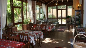 Un comedor con paredes de ladrillo, vigas de madera y ventanas grandes. Las mesas están cubiertas con manteles florales y sillas dispuestas alrededor.