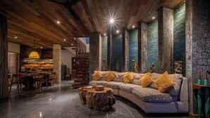 A modern living room with a wooden ceiling, a sectional sofa with patterned cushions, a round wooden coffee table, and a dining area with a wooden table and chairs. - Aryaswara Villa Ubud (Ubud)