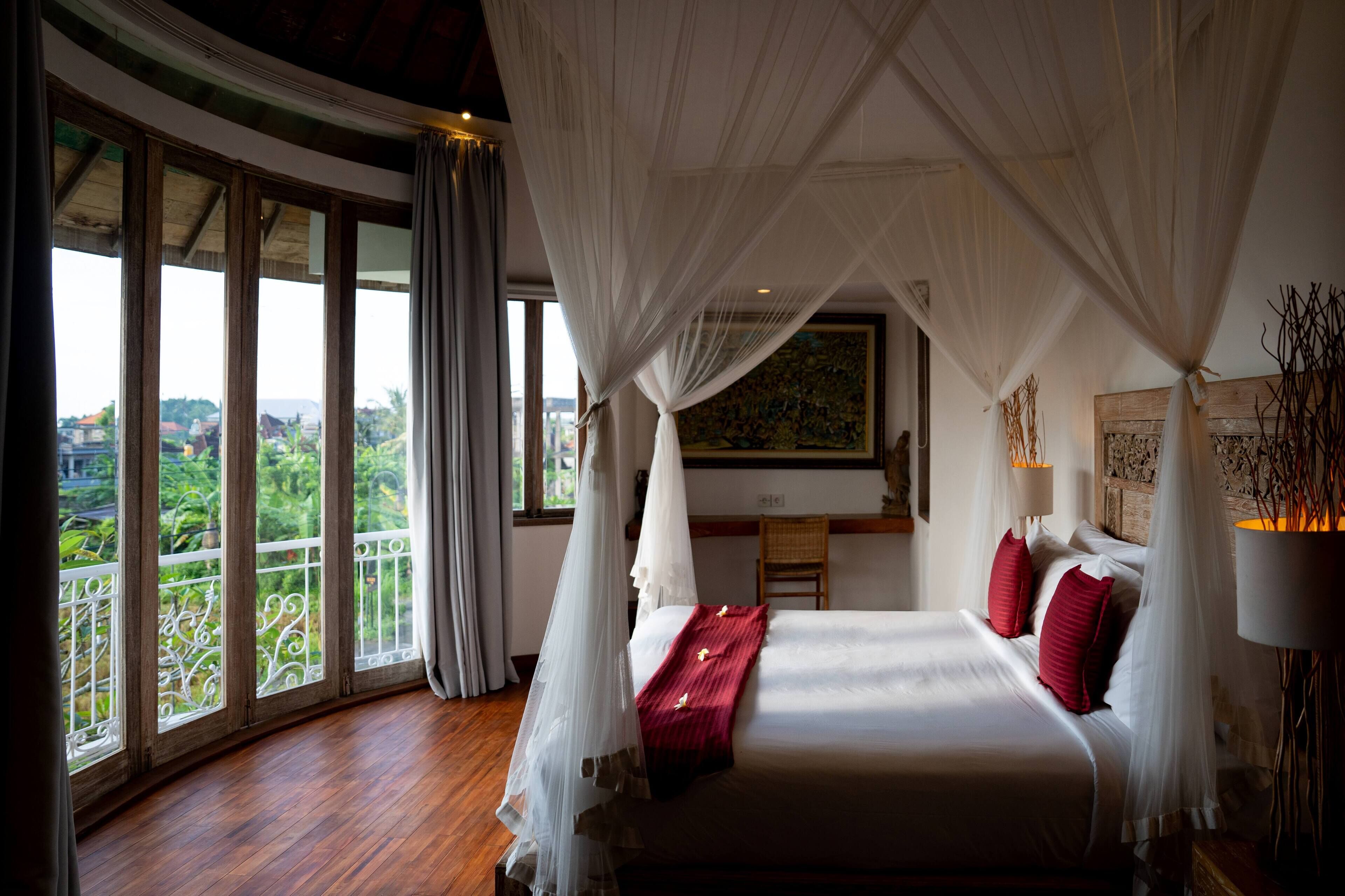 A bedroom with a large bed, white canopy, and a view of greenery through large windows.