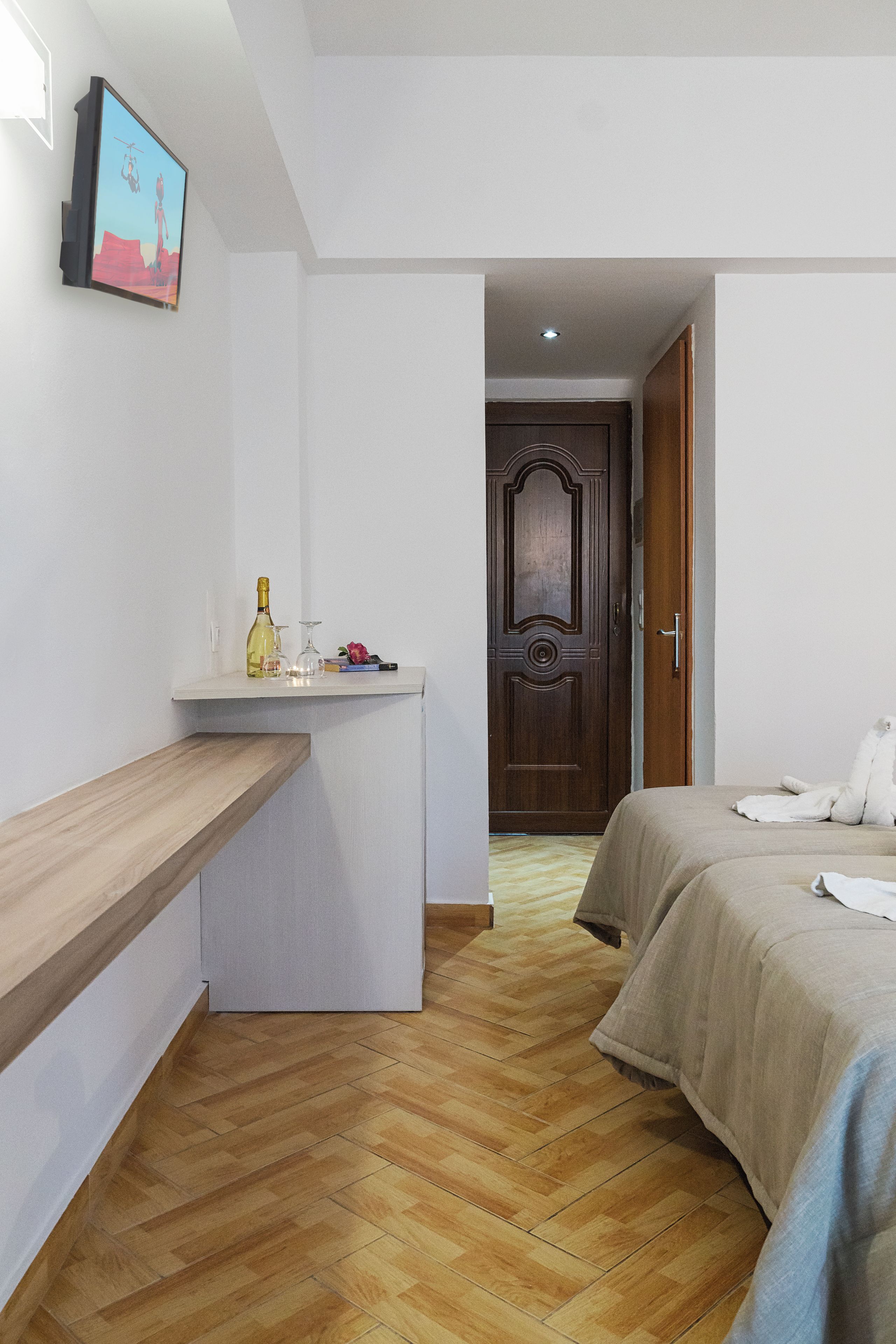 A hotel room with a wooden bench, a small table with a bottle and glasses, a bed with a blanket, and a wooden door in the background.