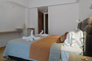Una habitación de hotel con una cama, una mesita de noche con una botella y vasos, un banco de madera y una puerta que da a otra habitación.