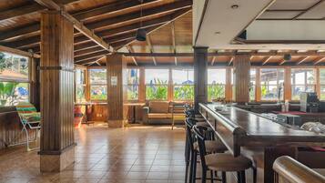 A spacious indoor area with wooden beams, a bar area with stools, and large windows offering views of greenery.