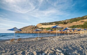 A beach with rows of sun loungers and umbrellas, rocky terrain, and a hilly backdrop. - Begeti Bay (Rethymno)