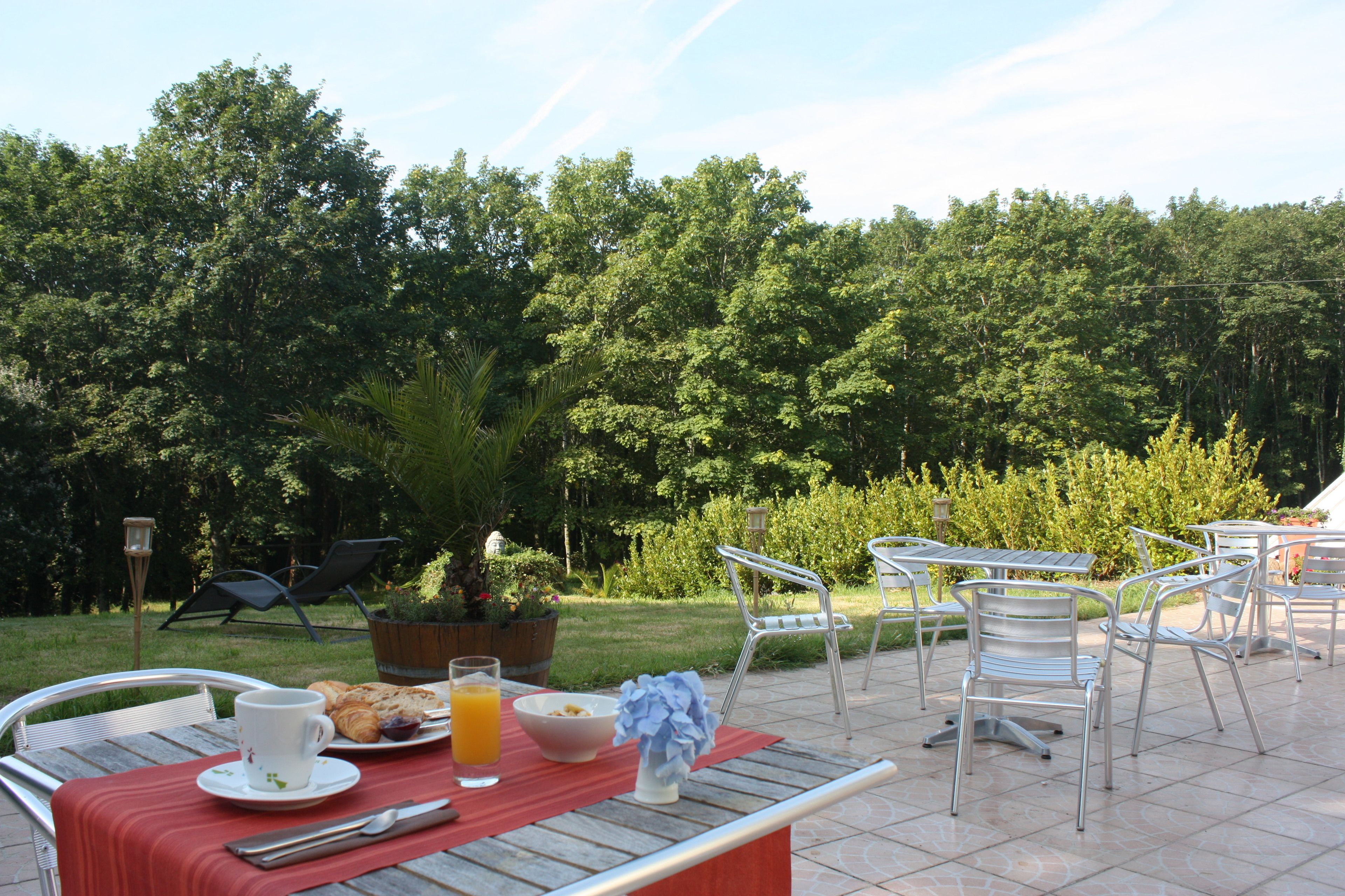 Een eethoek buiten met metalen stoelen en tafels, een houten tafel met een rode tafelkleed, een witte beker, een glas sinaasappelsap en een schaal eten.