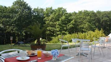 Een eethoek buiten met metalen stoelen en tafels, een houten tafel met een rode tafelkleed, een witte beker, een glas sinaasappelsap en een schaal eten.