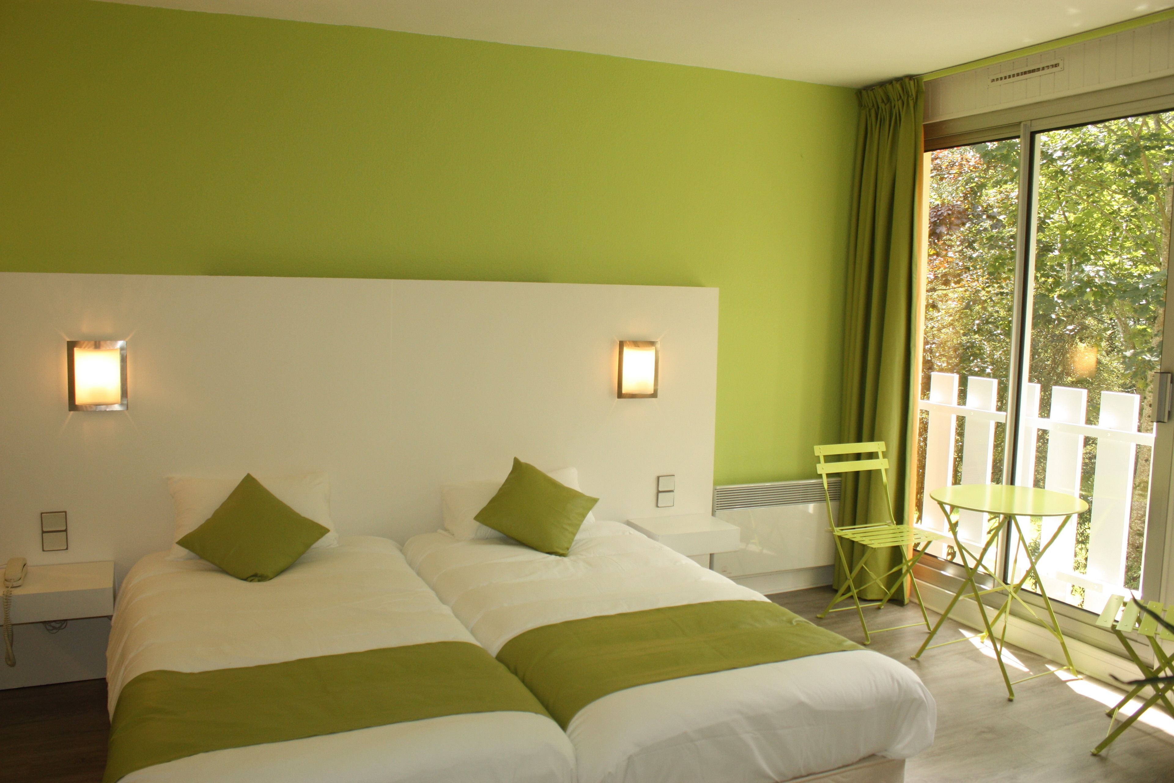 A hotel room with two beds, a small table, a chair, and a balcony with a view of trees.