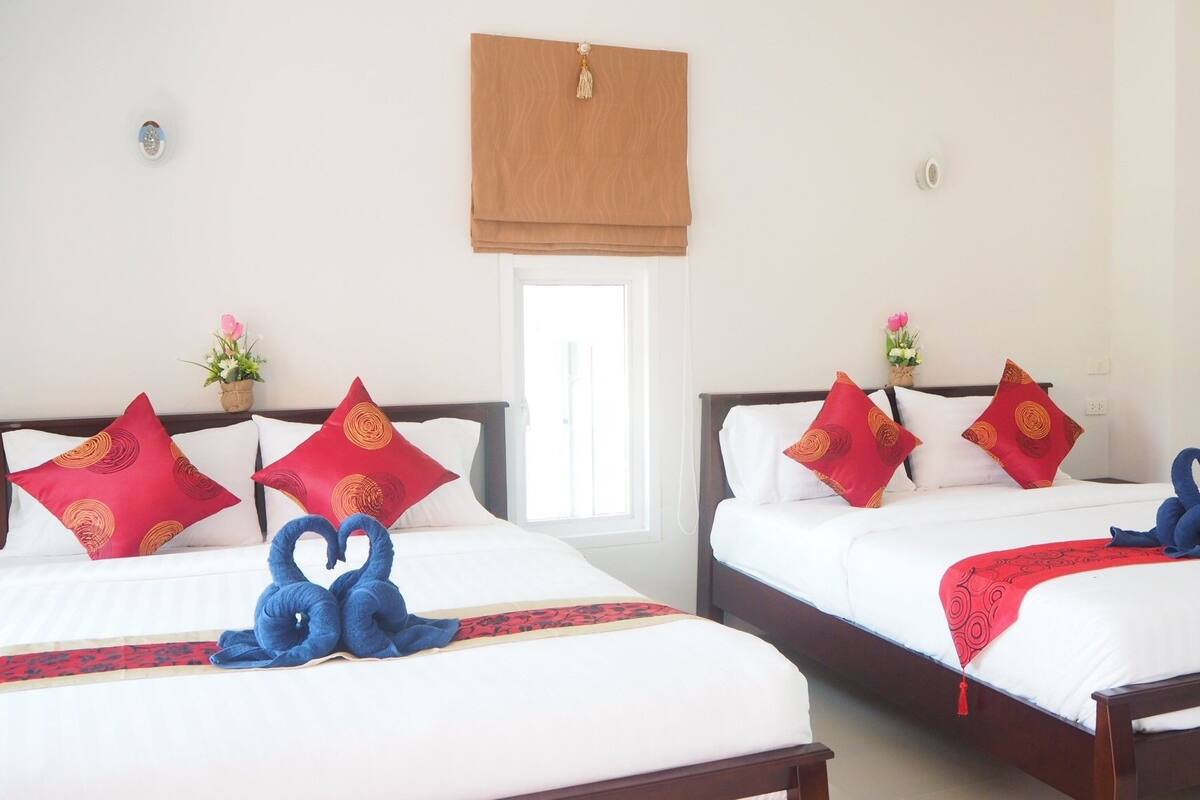 Ein Hotelzimmer mit zwei Betten, jeweils mit weißer Bettwäsche und roten Kissen mit einem kreisförmigen Muster. Auf den Betten liegen blaue Handtücher, und über dem Fenster ist ein brauner Jalousiewinkel angebracht.