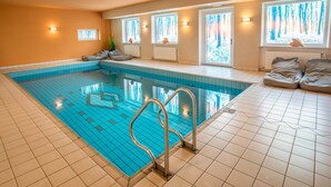 Una piscina interior con baldosas azules cristalinas, suelo de baldosas y un ventanal con vistas a árboles.