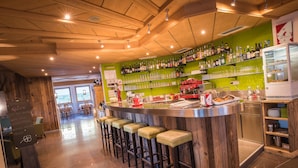 Zona de bar con taburetes de madera, una barra con vasos y botellas, y una pared verde con estantes llenos de botellas.