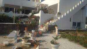 A modern white building with a staircase leading to a balcony. There are multiple potted plants and a stone wall in the foreground. - Kohili Studios (Paros)