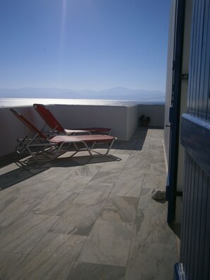 A balcony with two red lounge chairs and a table, overlooking a body of water and distant mountains. - Kohili Studios (Paros)