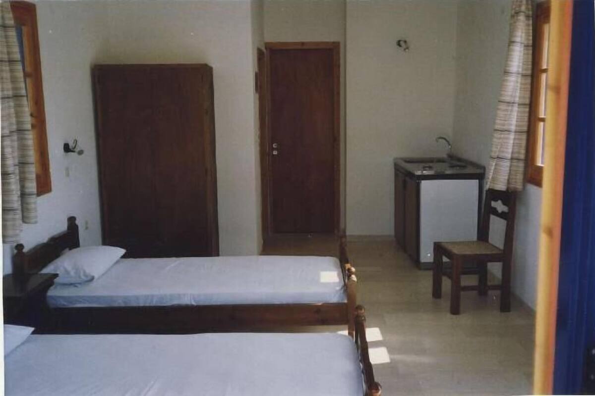 A small room with two single beds, a wooden wardrobe, a small desk with a chair, and a door leading to another room.