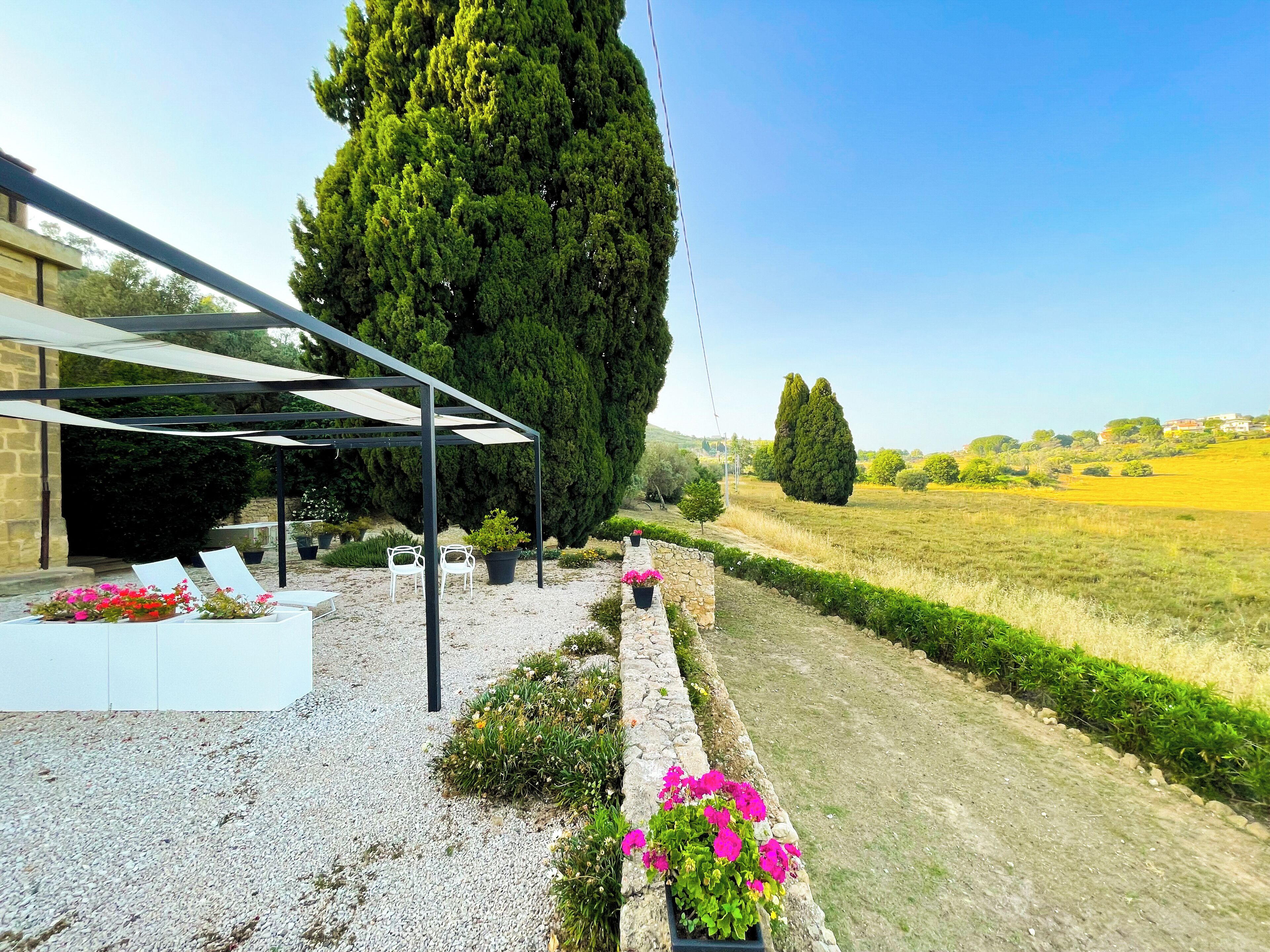 Un área de estar al aire libre con techo, sillas blancas y macetas con flores.