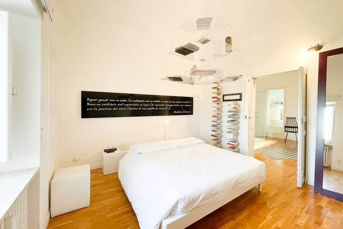 A bedroom with a bed, a bedside table, a wall-mounted shelf with books, and a hanging light fixture.
