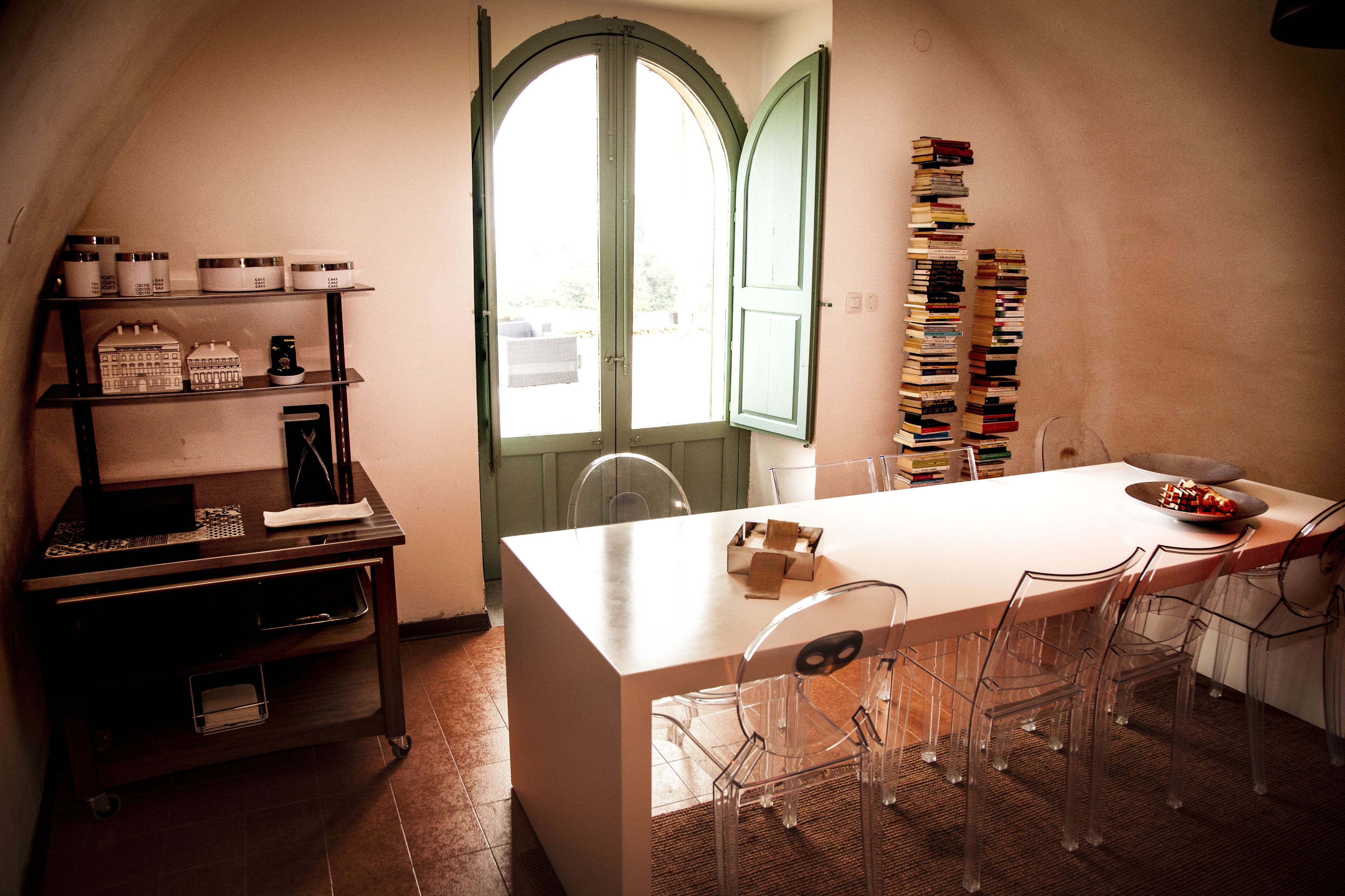 A dining area with a white table, clear chairs, a wooden sideboard with jars, and a stack of books against the wall.