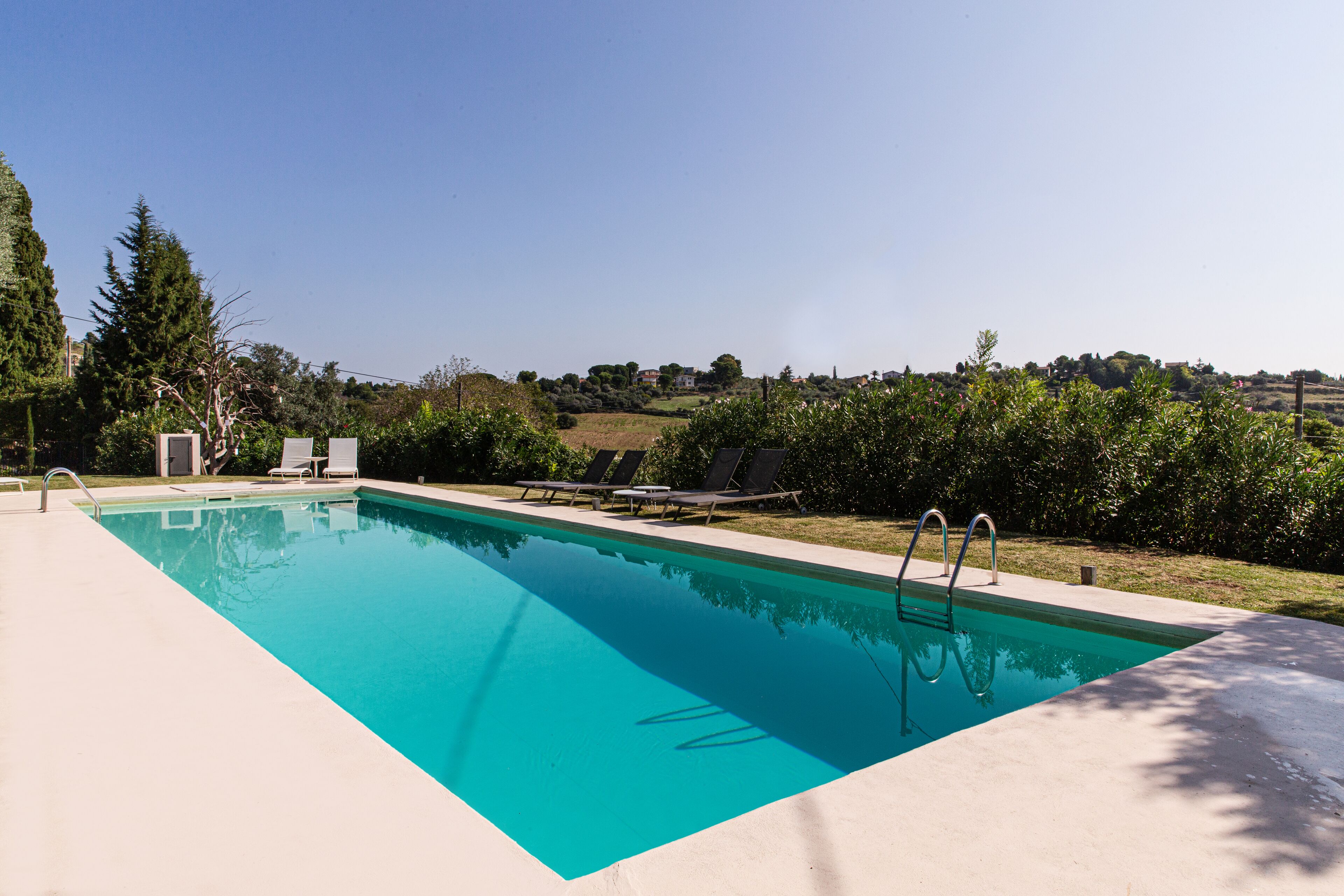 A rectangular swimming pool with a clear blue water surface, surrounded by a concrete deck. There are lounge chairs and a small table near the pool's edge.