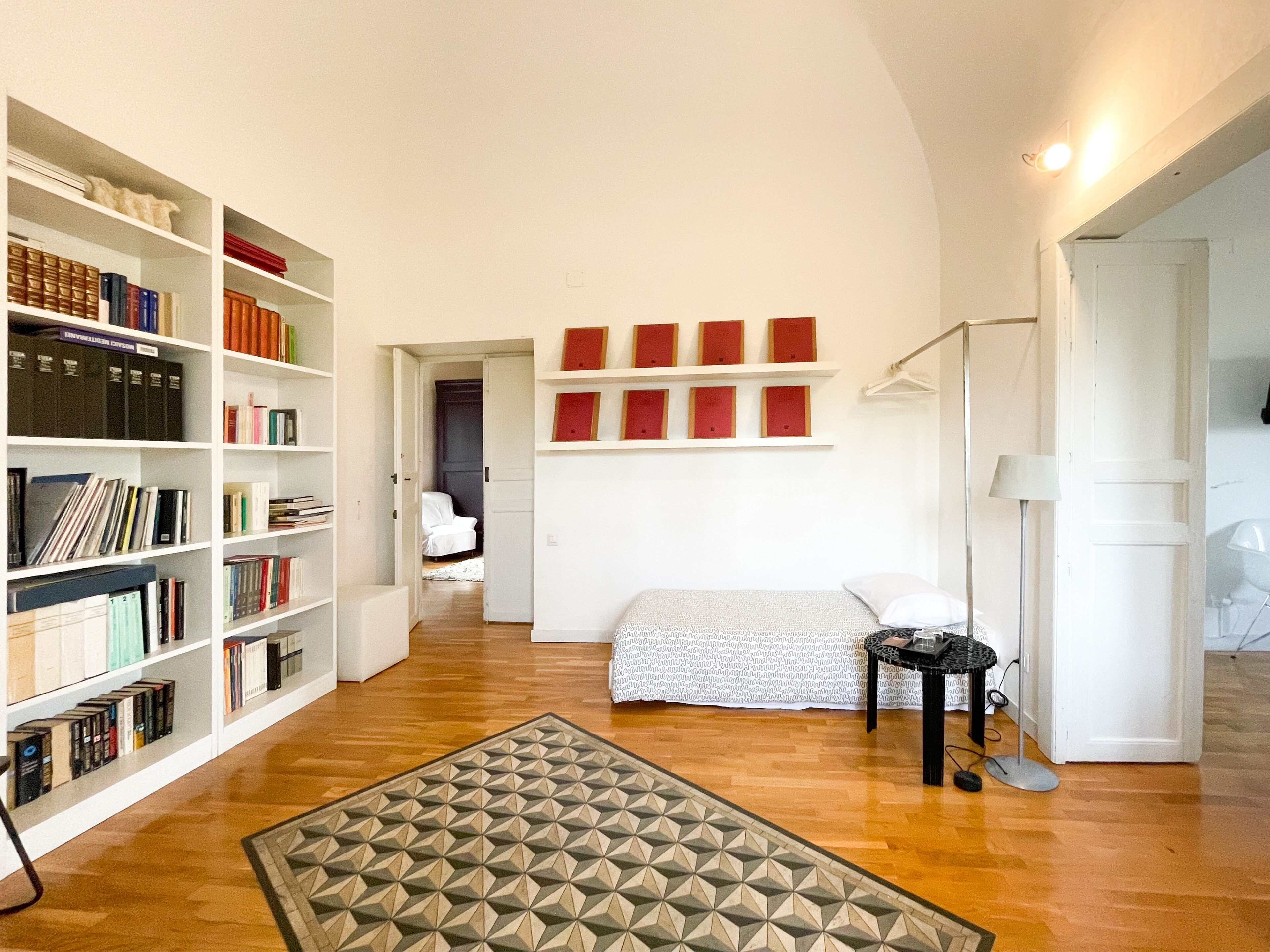 A spacious room with a large bookshelf, a bed with a patterned bedspread, and a small table with a lamp.