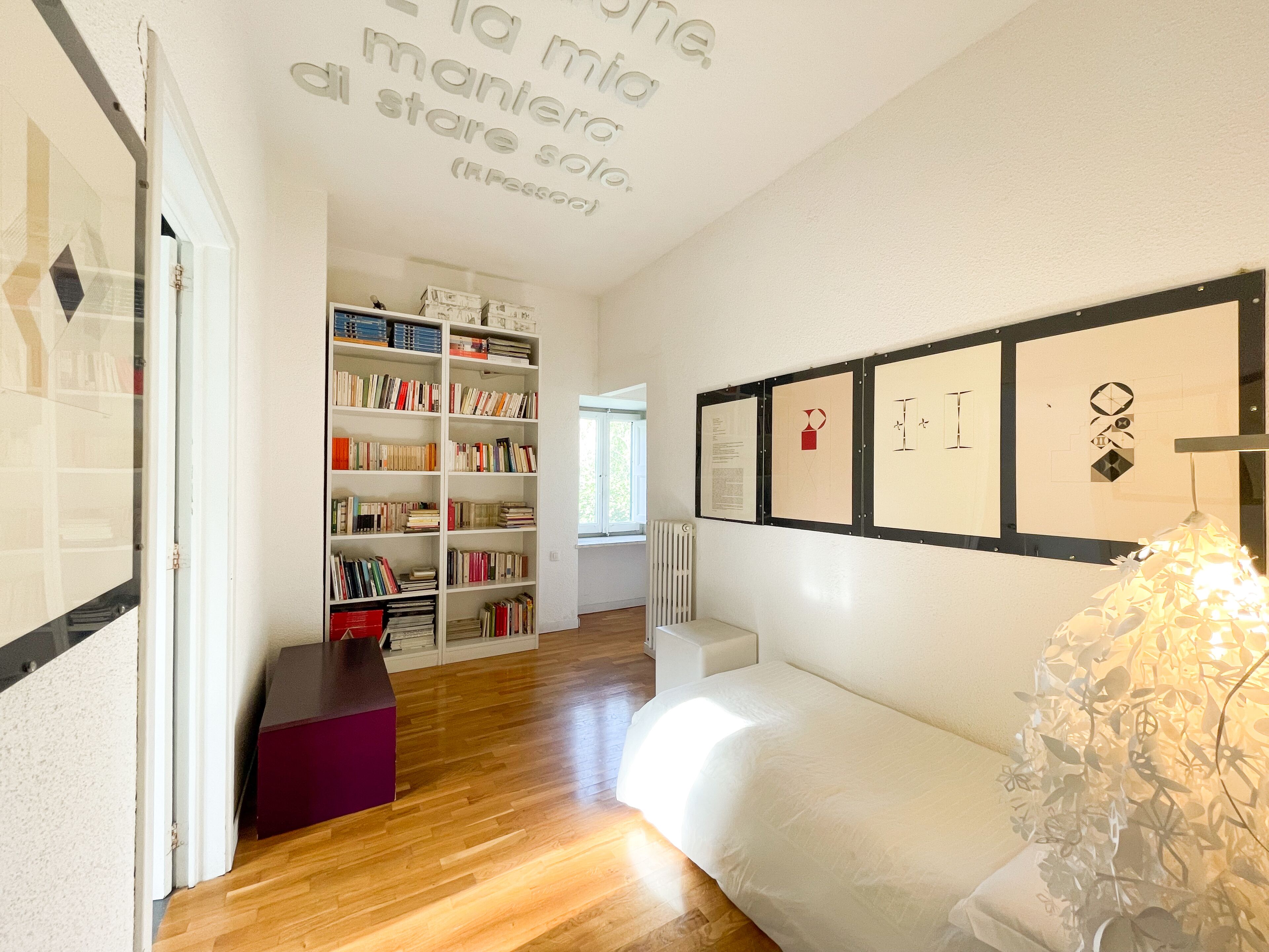 A room with a wooden floor, a white bed, a bookshelf, and framed artwork on the wall.