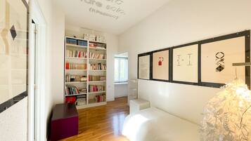 A room with a wooden floor, a white bed, a bookshelf, and framed artwork on the wall.