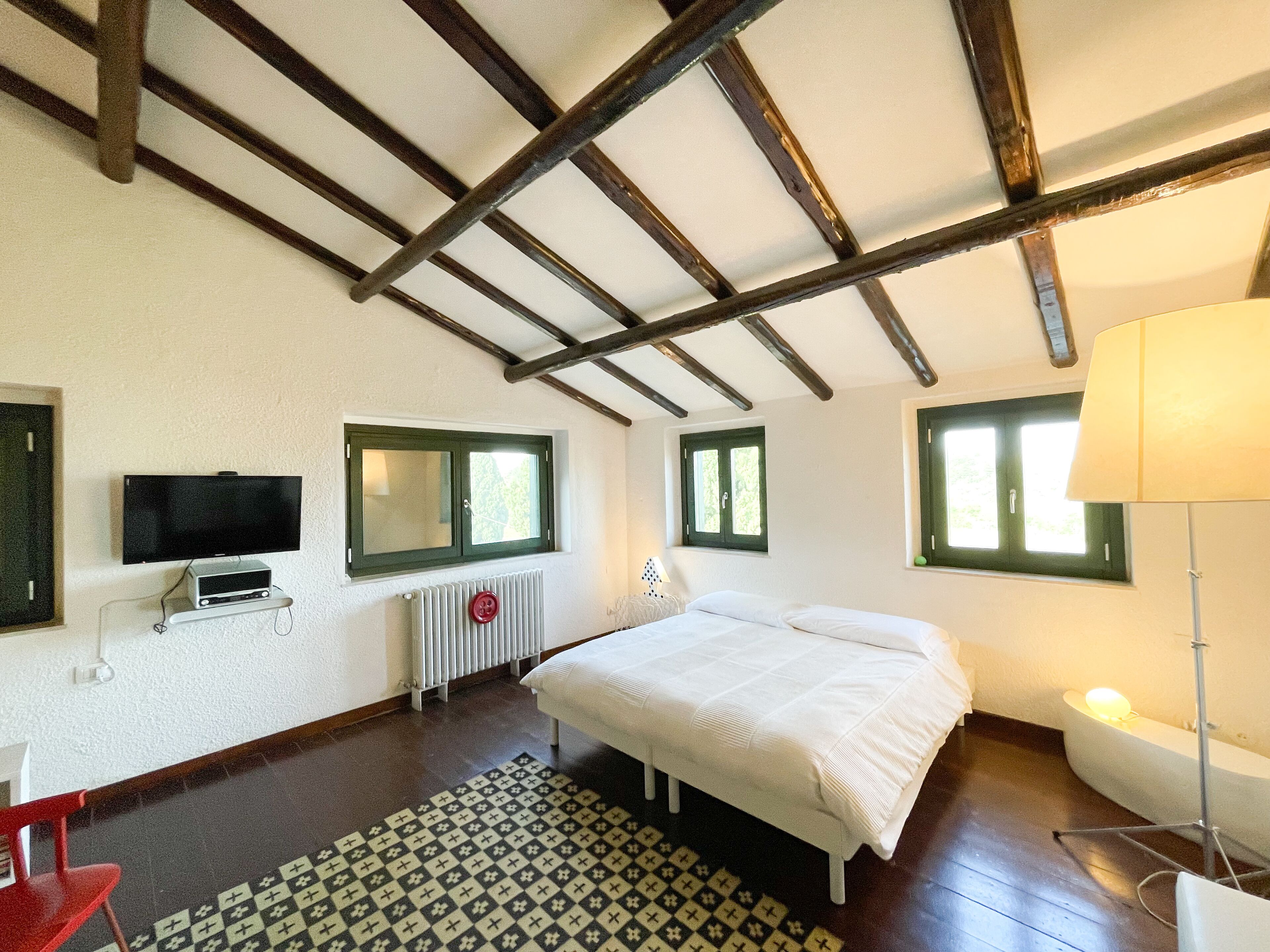 Un dormitorio con cama, televisión, radiador y dos ventanas.