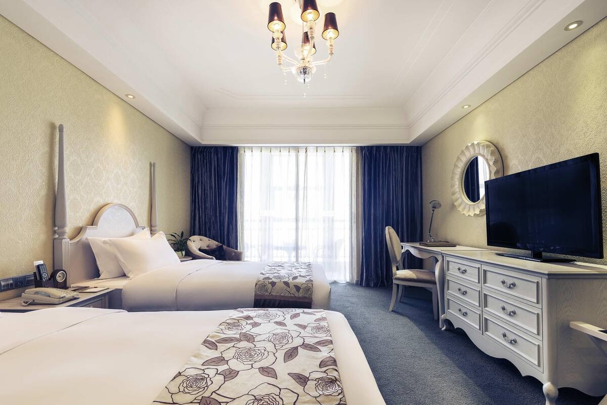 Habitación de hotel con dos camas, un escritorio, una silla, un televisor y un ventanal con cortinas.