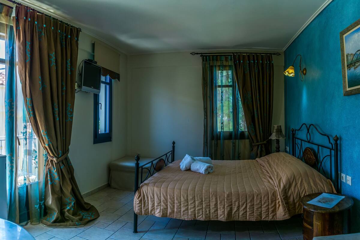 Un dormitorio con cama, televisión, ventana con cortinas, lámpara y un cuadro en la pared.