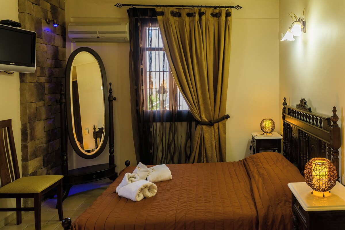A hotel room with a bed, a chair, a mirror, a window with curtains, a television, and a nightstand with a lamp.
