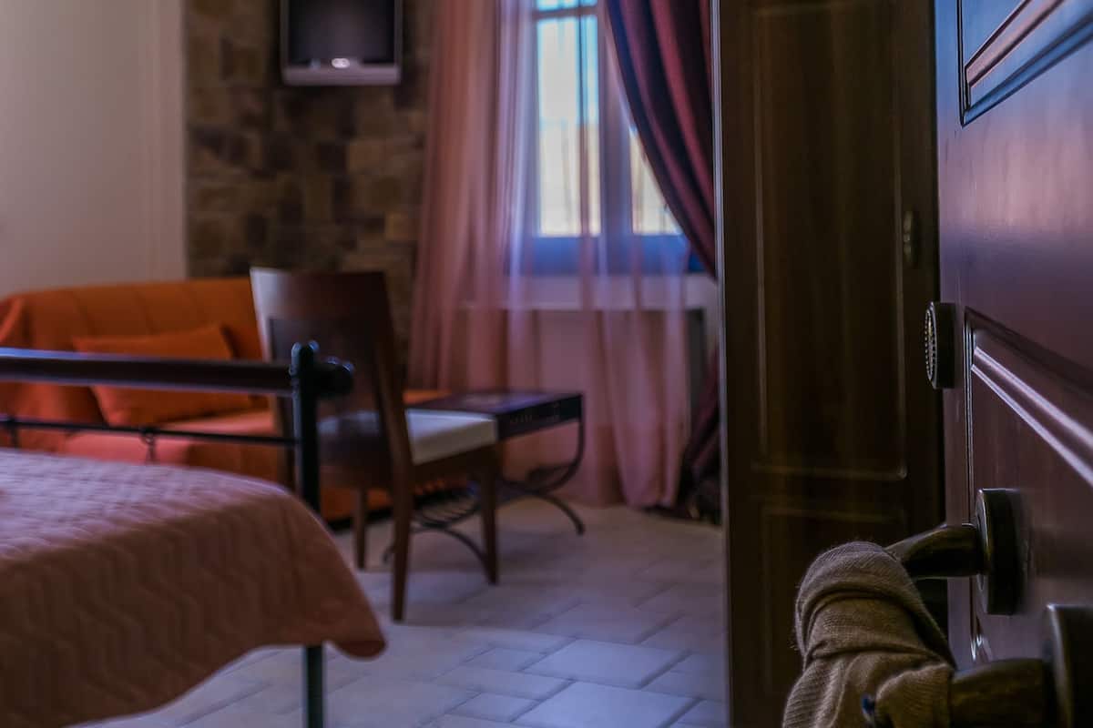 Una habitación con una cama, una silla, un televisor montado en una pared de piedra, una puerta de madera y una ventana con cortinas.