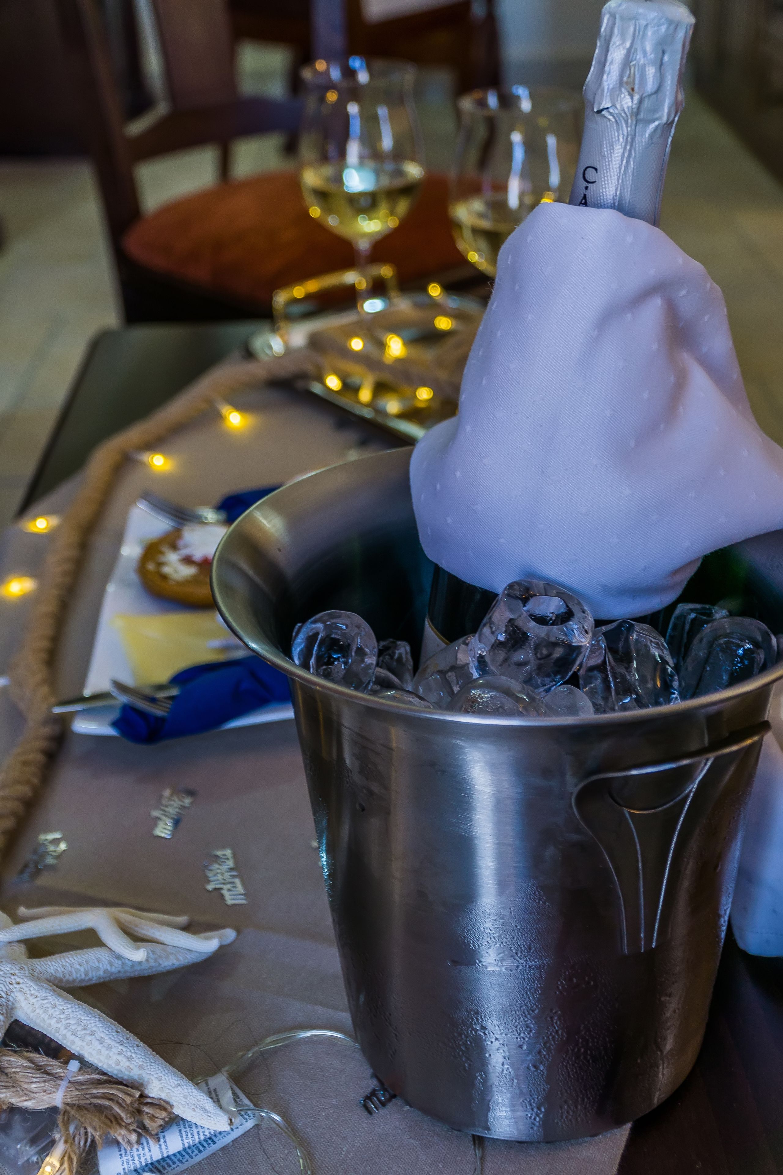 A metal bucket filled with ice cubes, a bottle of champagne, and a glass of white wine on a table with a placemat.