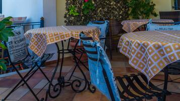 Un espace patio avec des tables recouvertes de nappes à motifs, une chaise sur laquelle est posée une veste en jean, et un mur décoratif agrémenté de plantes et d’une rose.