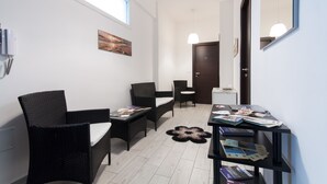 Ein modernes Zimmer mit einem großen Fenster, einem schwarzen Rattan-Sofa, einem kleinen Tisch mit Zeitschriften und einem schwarzen Regal mit Büchern.