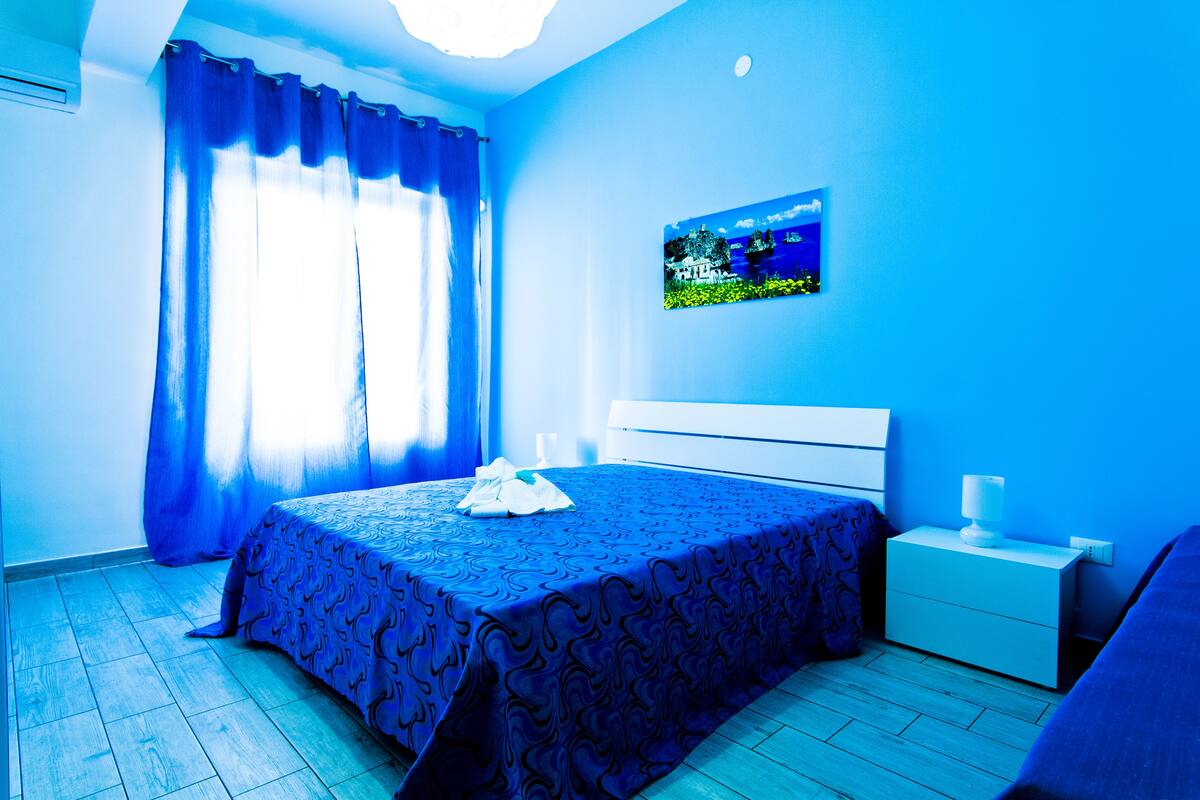 Een slaapkamer met een bed, nachtkastje, een aan de muur bevestigde televisie en een raam met gordijnen.