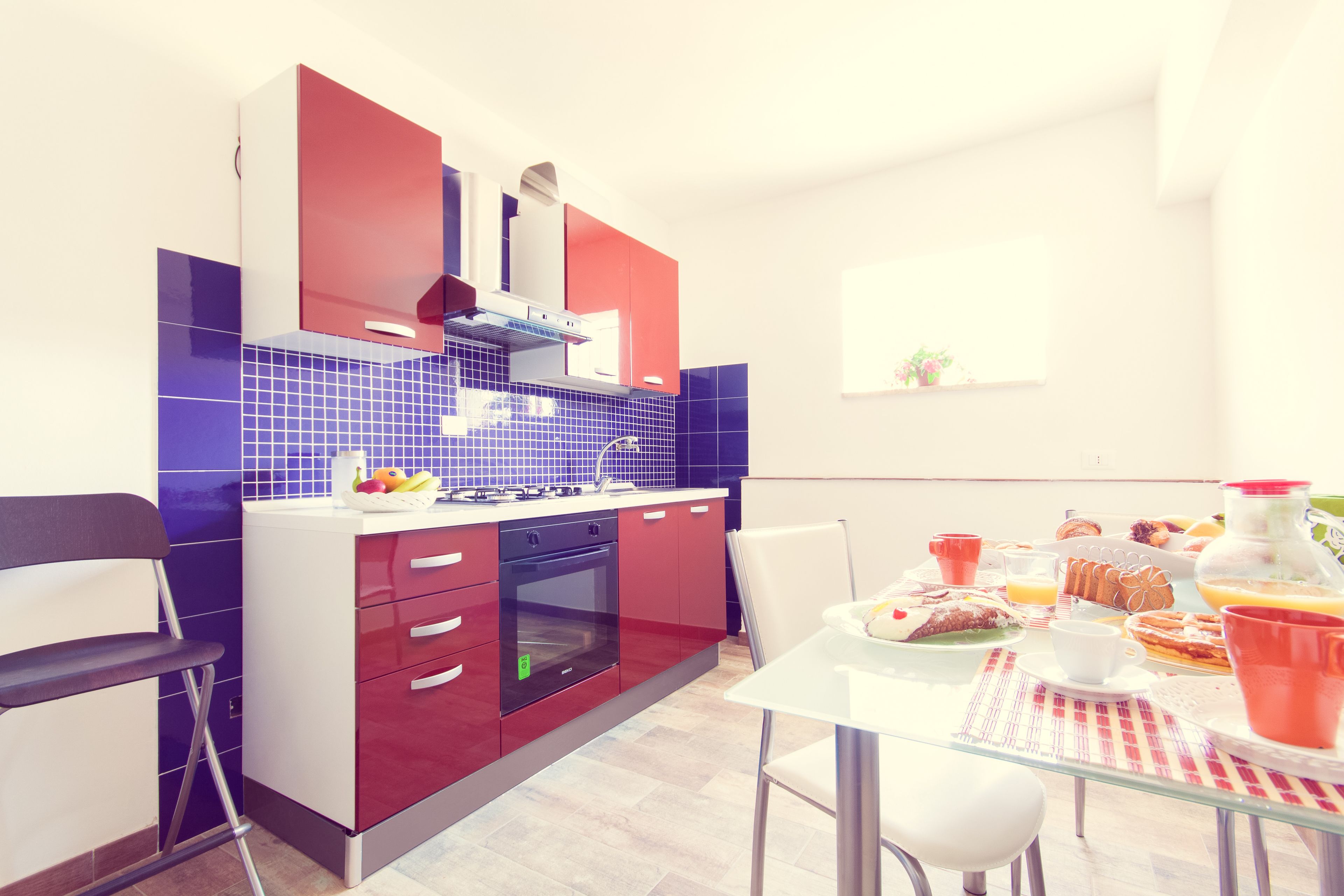 Una cocina moderna con armarios rojos y blancos, un salpicadero de azulejos azules y un comedor con mesa y sillas de vidrio.