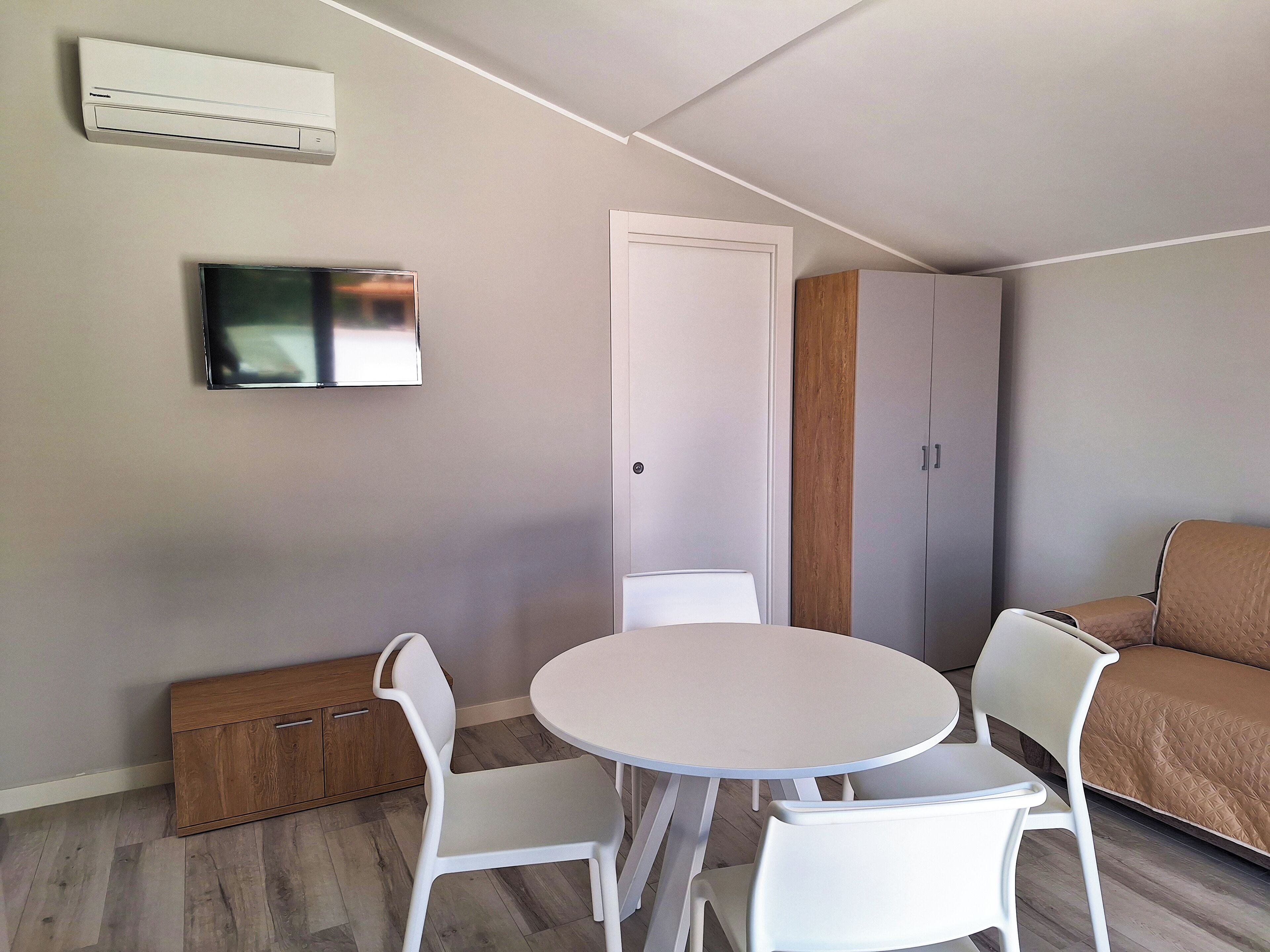 Ein kleiner Wohnraum mit rundem Tisch und zwei Stühlen, einem an der Wand befestigten Fernseher, einem Kleiderschrank und einer Tür.