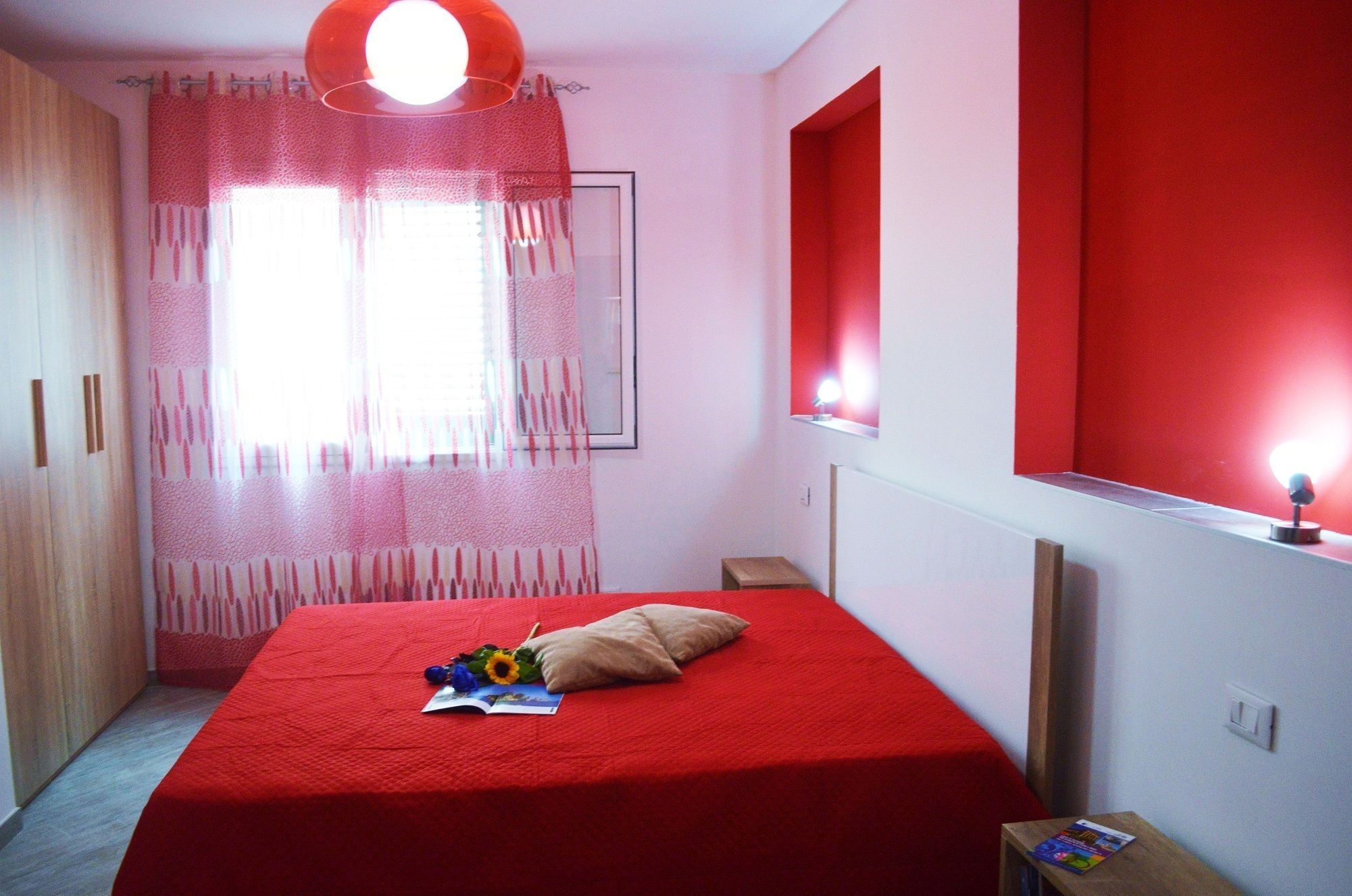 Een kamer met een rood bed, een raam met dunne gordijnen, een nachtkastje met een lamp en een rode accentmuur.
