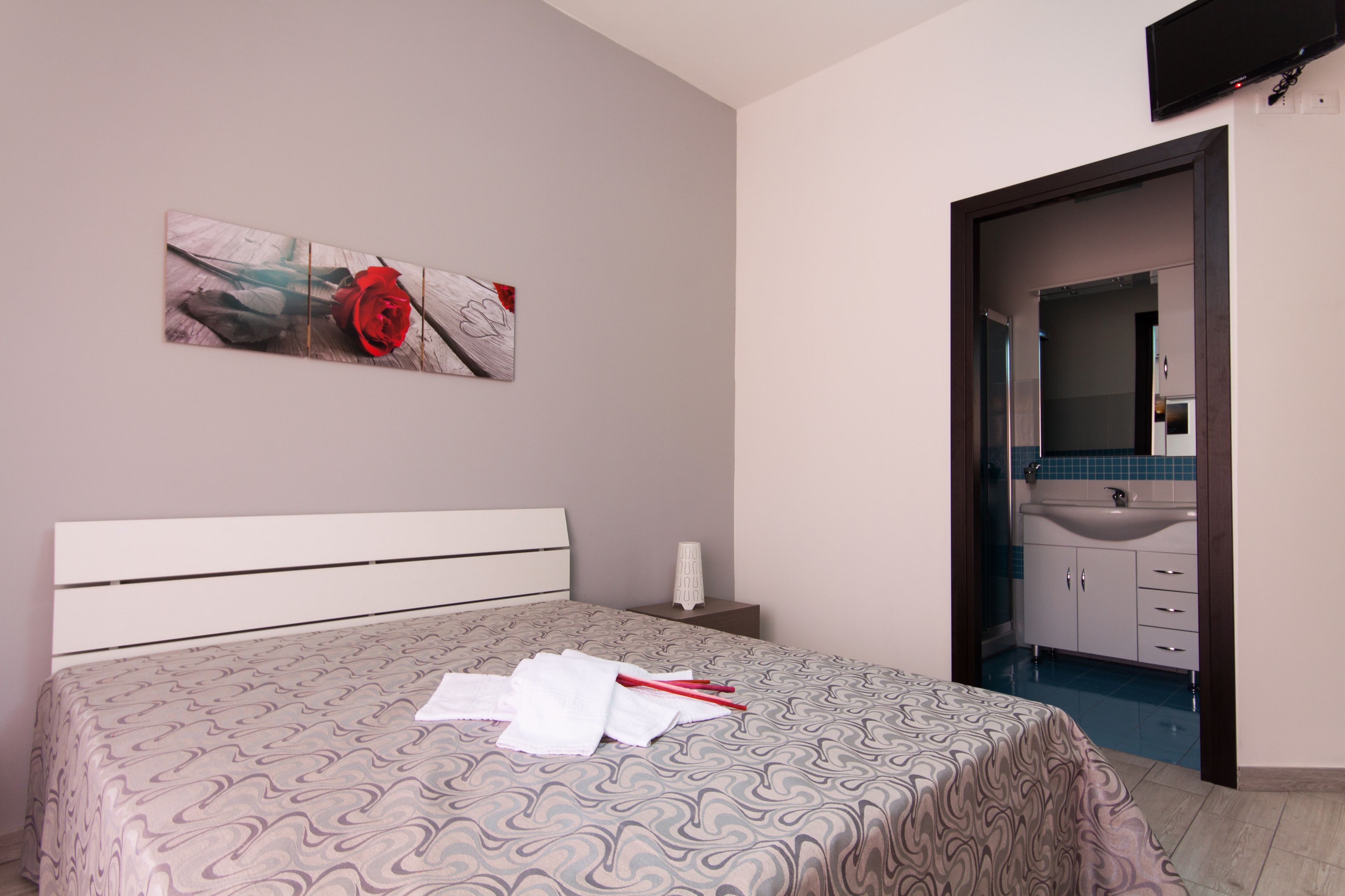 Une chambre à coucher avec une couvre-lit à motifs, une œuvre d’art représentant une rose rouge accrochée au mur, une table de chevet avec une lampe, et une vue sur une salle de bain équipée d’un lavabo et de meubles de rangement.