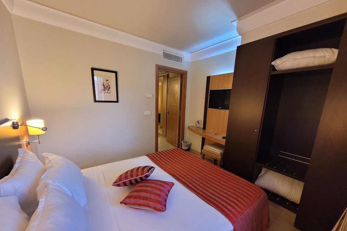 A hotel room with a bed, a bedside lamp, a framed picture on the wall, a wooden wardrobe, and a small table with a lamp.