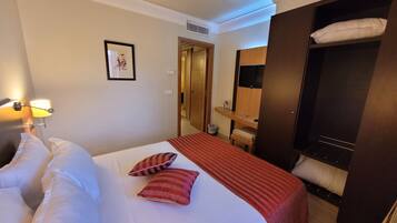 Habitación de hotel con cama, lámpara de noche, un cuadro enmarcado en la pared, un armario de madera y una mesita con lámpara.