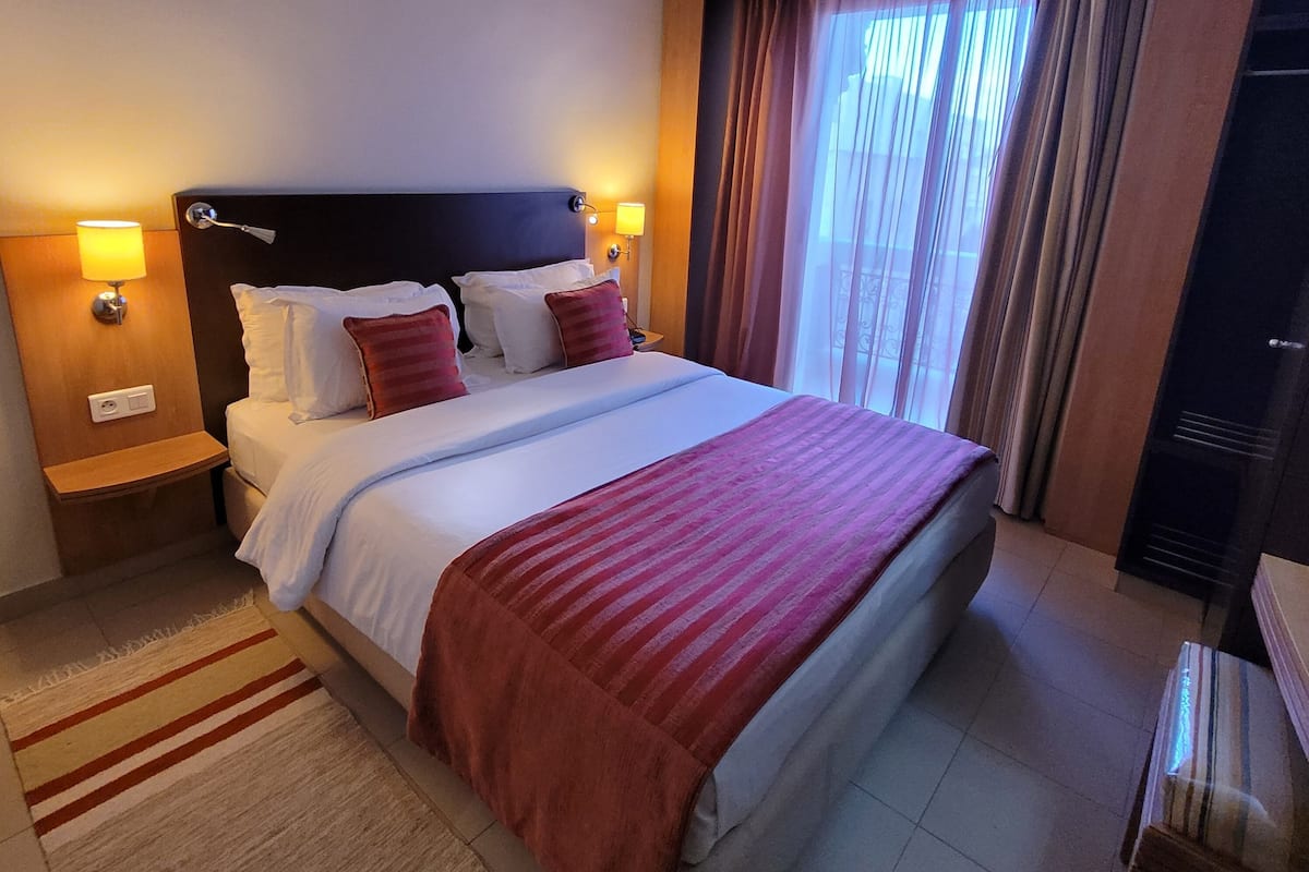 A hotel room with a large bed, two bedside tables with lamps, a wooden headboard, a window with curtains, and a small table with a chair.