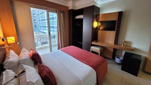Ein Hotelzimmer mit Bett, Fernseher, Schreibtisch und einem Balkon mit Blick auf Gebäude.