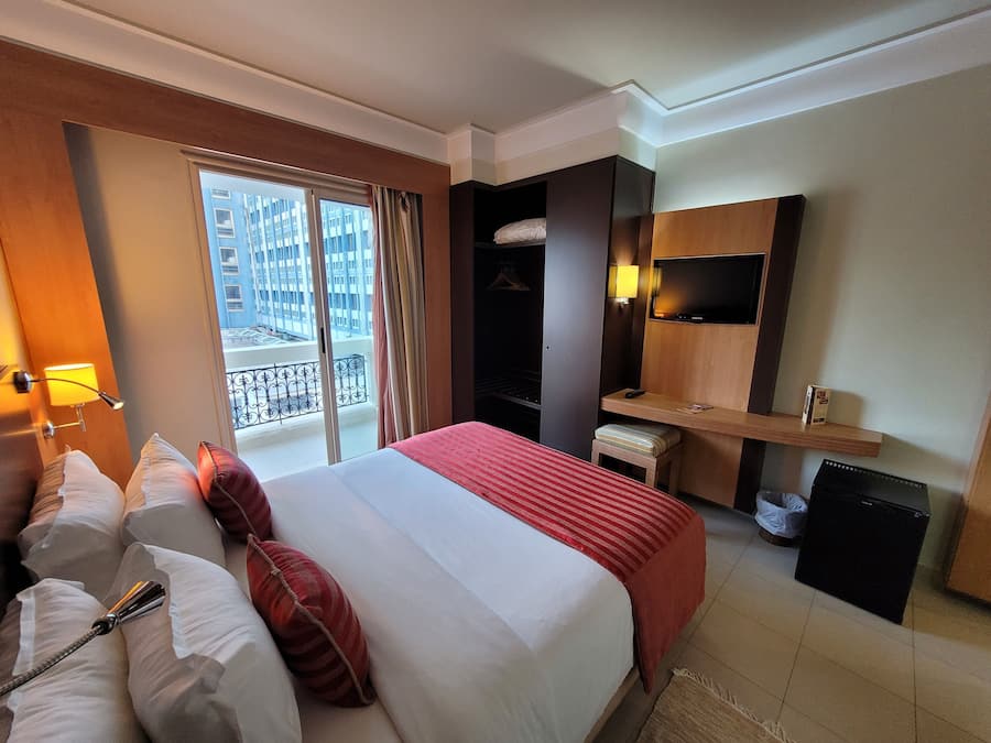 A hotel room with a bed, a TV, a desk, and a balcony with a view of buildings.