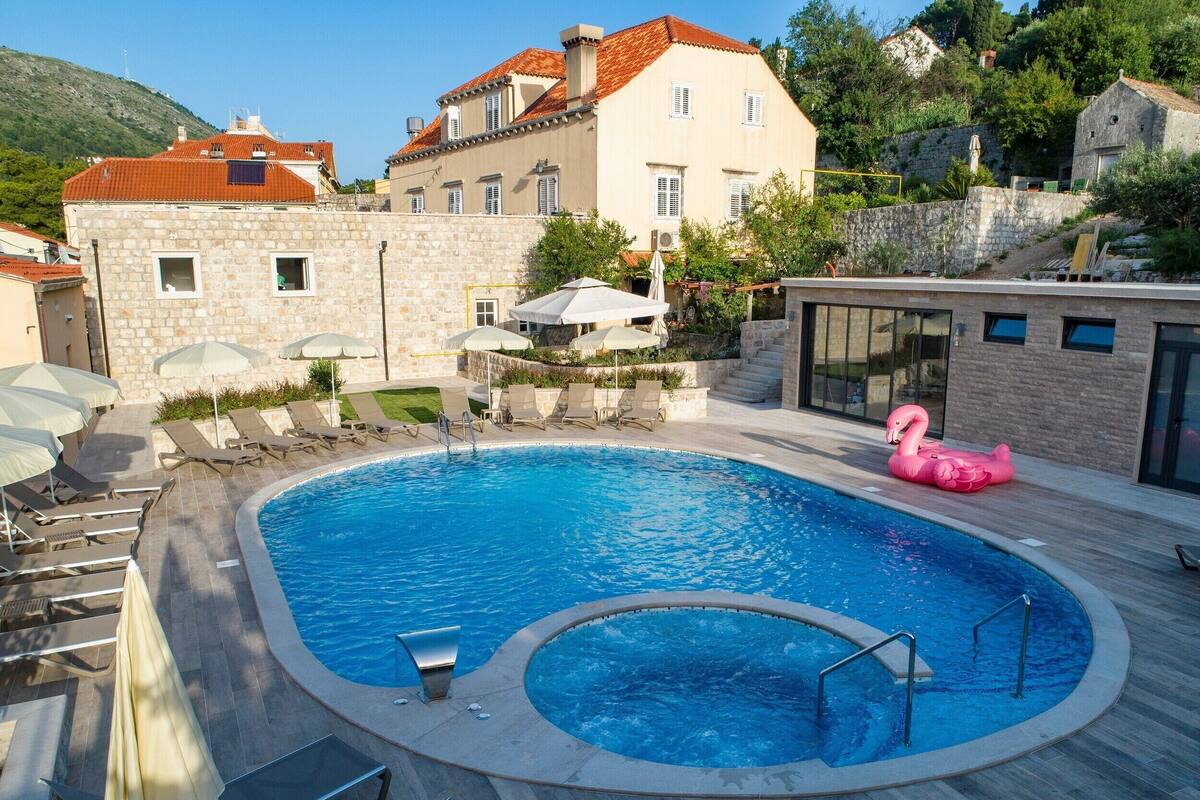 A swimming pool with lounge chairs and umbrellas, surrounded by stone buildings and a hillside.