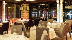 Un restaurante con techos altos y vigas a la vista, pilares de piedra y una araña. Las mesas están dispuestas con manteles blancos y sillas.