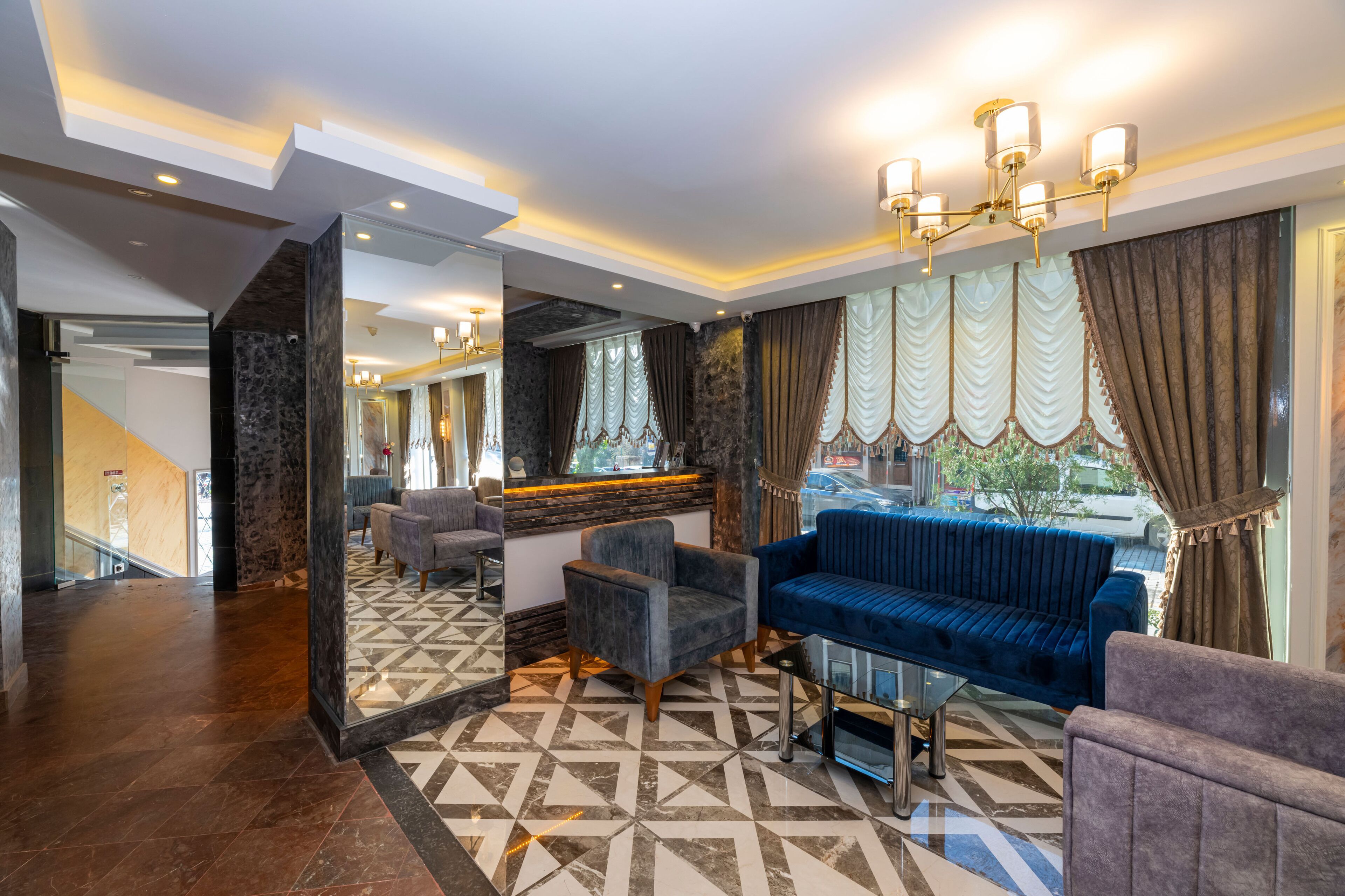 A modern lobby with a blue sofa, armchairs, and a glass coffee table. A mirror reflects the space, and there's a staircase in the background.