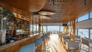 Un bar moderno con techo de madera y taburetes, amplios ventanales con vista a la ciudad y una barra repleta de botellas y vasos.